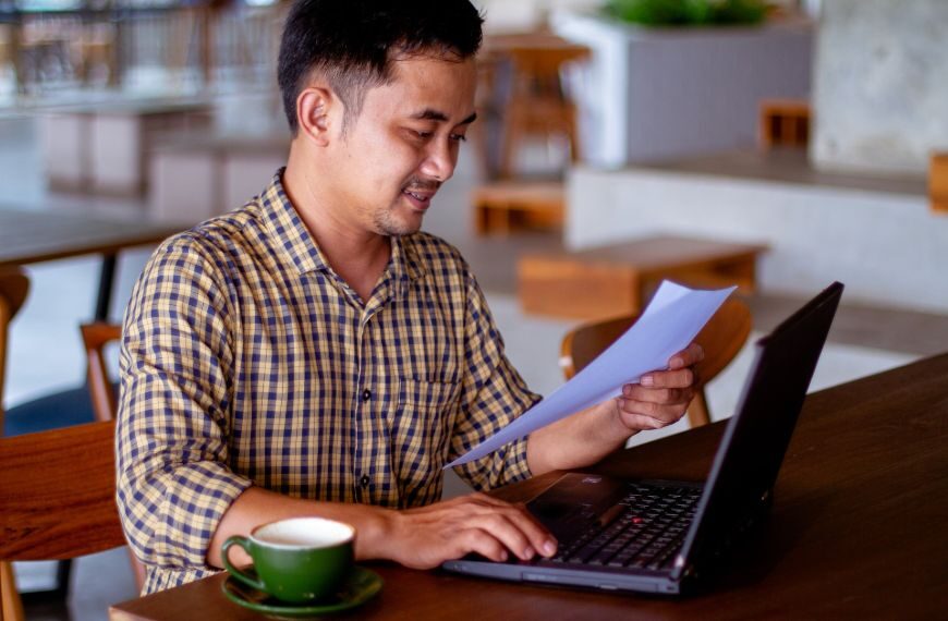 Man sitting at computer holding a piece of paper smiling. He is reading good new and is feeling happy.